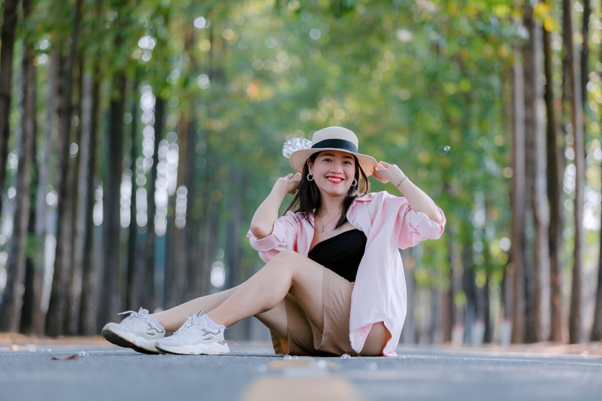 Smiling woman in casual attire sitting on a tree-lined road enjoying a sunny day.