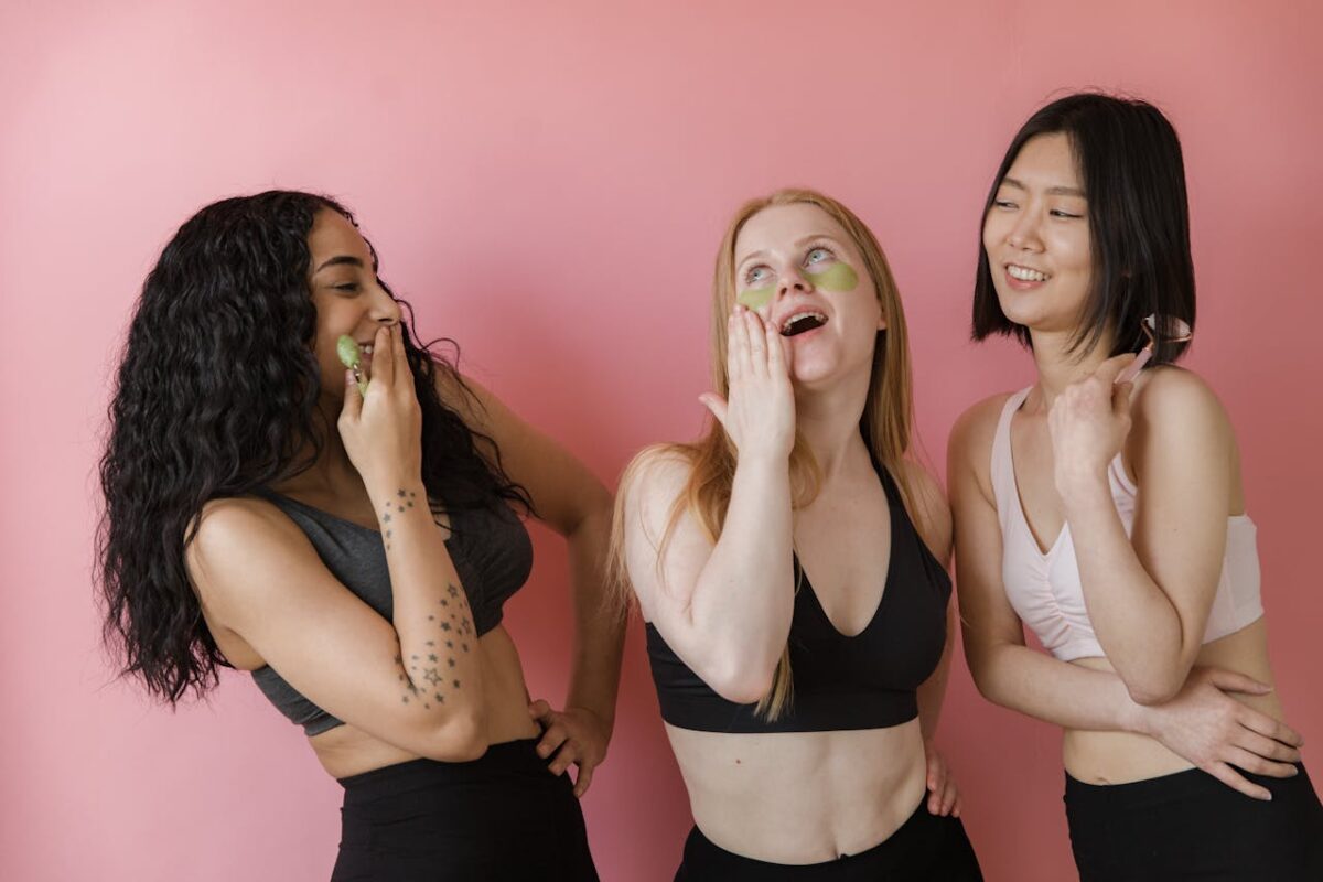 Three women enjoying a beauty session, applying facial masks with joy and laughter against a pink background.