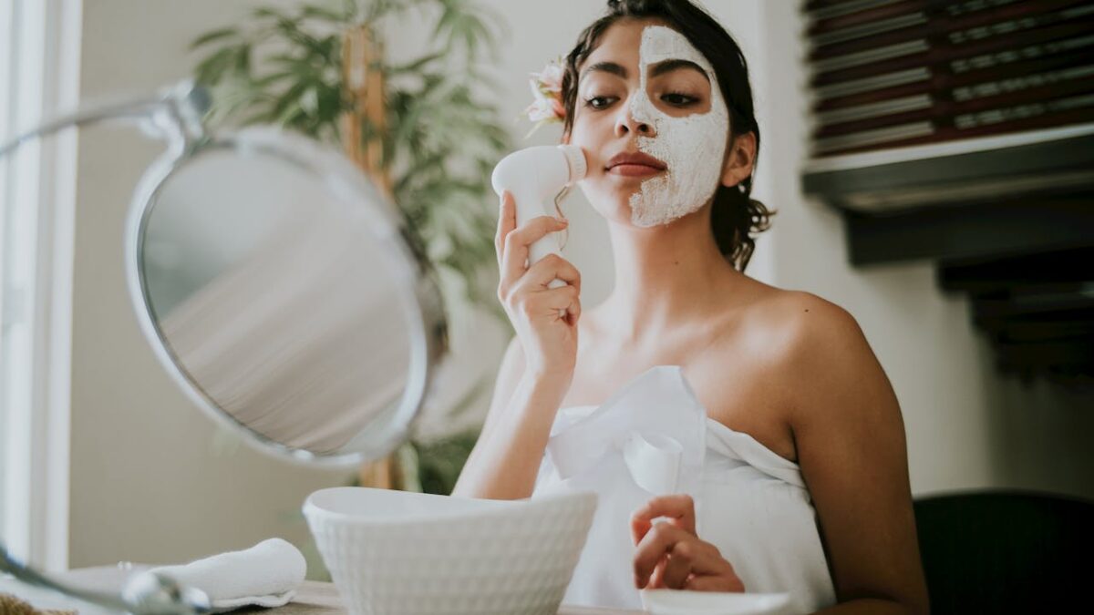 A woman uses a facial brush to apply a clay mask during a relaxing skincare routine.