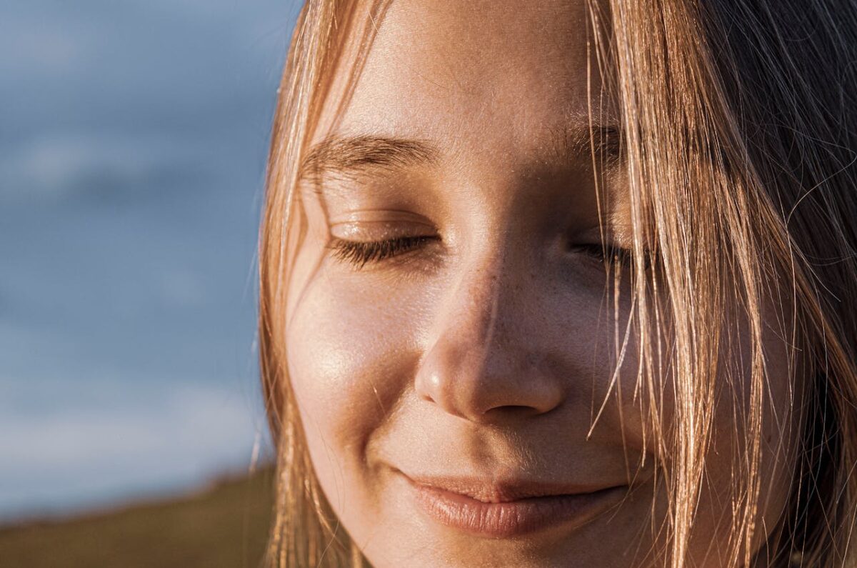 Close-up of a smiling woman with closed eyes, sunlit hair, and a serene expression outdoors.